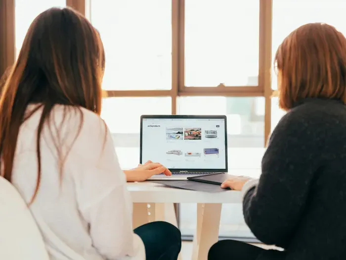 Two women sitting at a bright office table collaborating on a laptop, discussing website design project